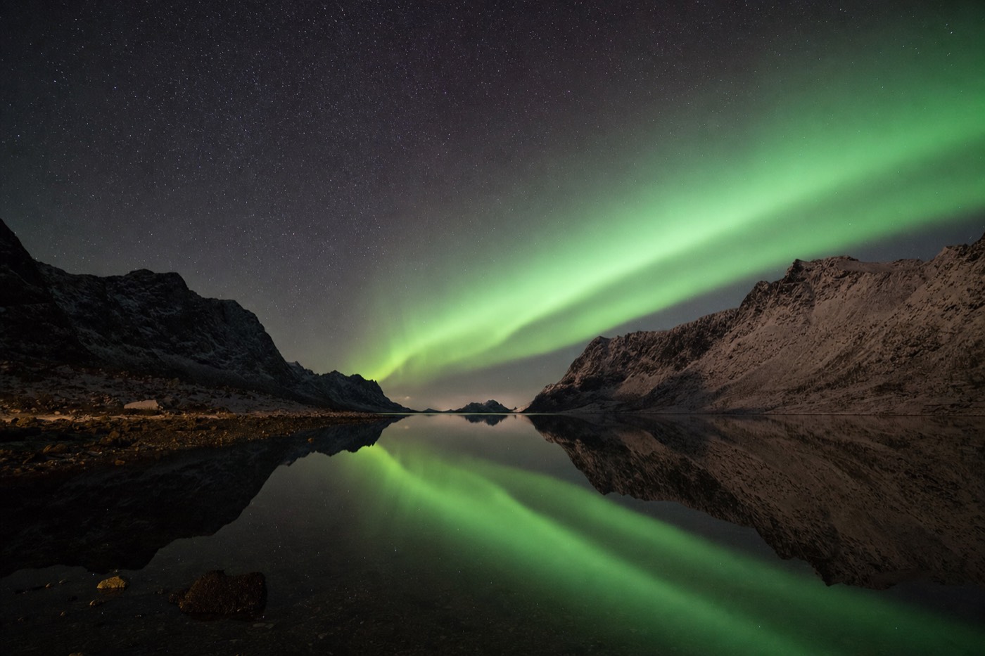 A ribbon of green aurora borealis mirrored in still black fjord water between mountain silhouettes