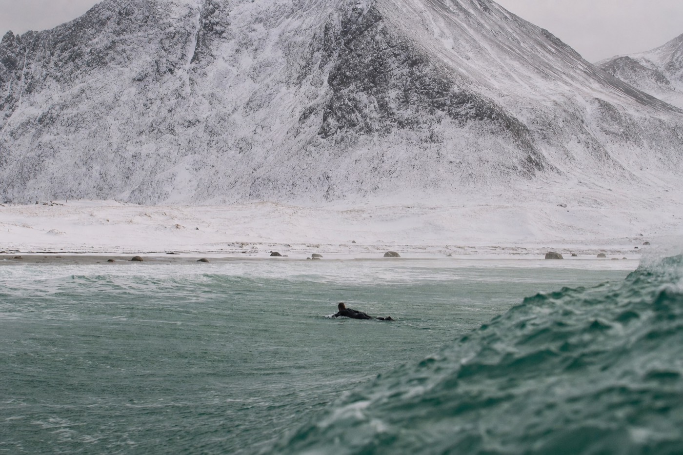 A surfer paddling out through cold Arctic waves at Unstad beach in Lofoten, mountains behind